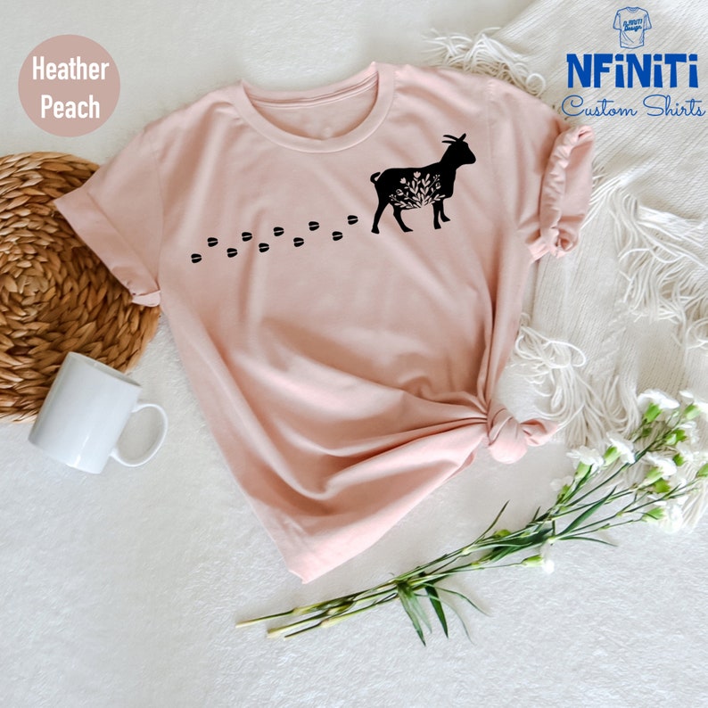 Pu&ograve; includere: Una t-shirt color pesca sfumato con una silhouette nera di una capra con fiori e foglie all'interno del suo corpo. La capra cammina verso destra e lascia una scia di impronte nere dietro di s&eacute;. Il testo "NFINITI Custom Shirts" &egrave; nell'angolo in alto a destra dell'immagine.