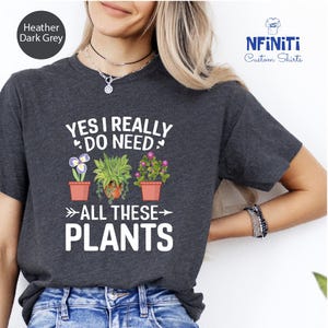 Può includere: Maglietta color grigio scuro melange con la scritta "YES I REALLY DO NEED ALL THESE PLANTS" e tre piante in vaso con fiori.