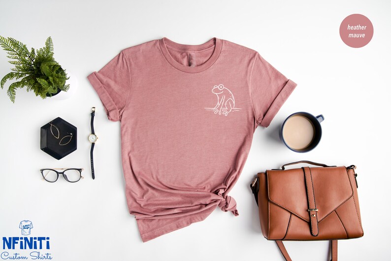 May include: A heather mauve t-shirt with a white outline of a frog on the front. The shirt is laid flat on a white surface with a mug, a purse, glasses, and a watch.