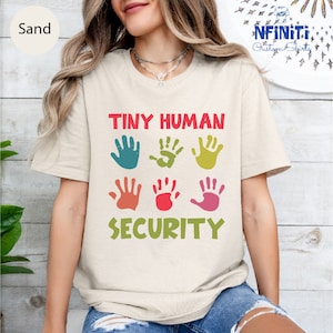 May include: A beige t-shirt with the text "Tiny Human Security" in colorful letters. The text is surrounded by six colorful handprints.