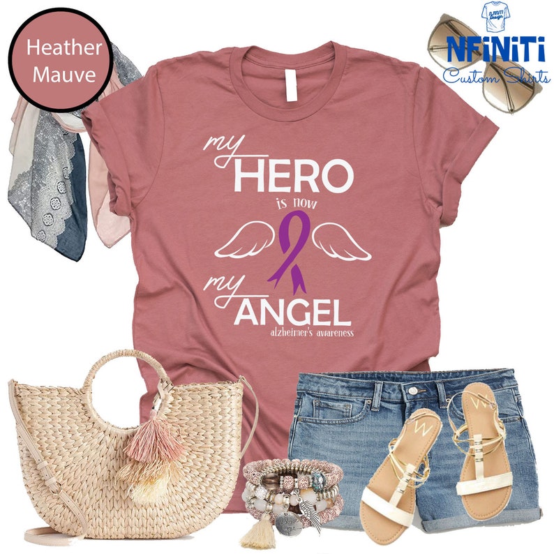 May include: Heather mauve t-shirt with the text "my HERO is now my ANGEL" and a purple ribbon, with the words "Alzheimer's awareness" below. The image also includes a straw tote bag, denim shorts, sandals, bracelets, and sunglasses.