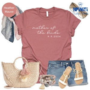 May include: A heather mauve t-shirt with the text "mother of the bride 9.9.2024" printed on it. The shirt is paired with denim shorts, a straw tote bag with pink tassels, and white sandals.