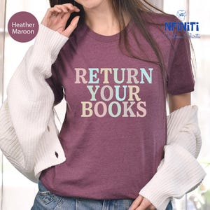 May include: Maroon t-shirt with the text "RETURN YOUR BOOKS" in pastel colors. The shirt is a heathered fabric and has short sleeves. The image also shows a white sweater.