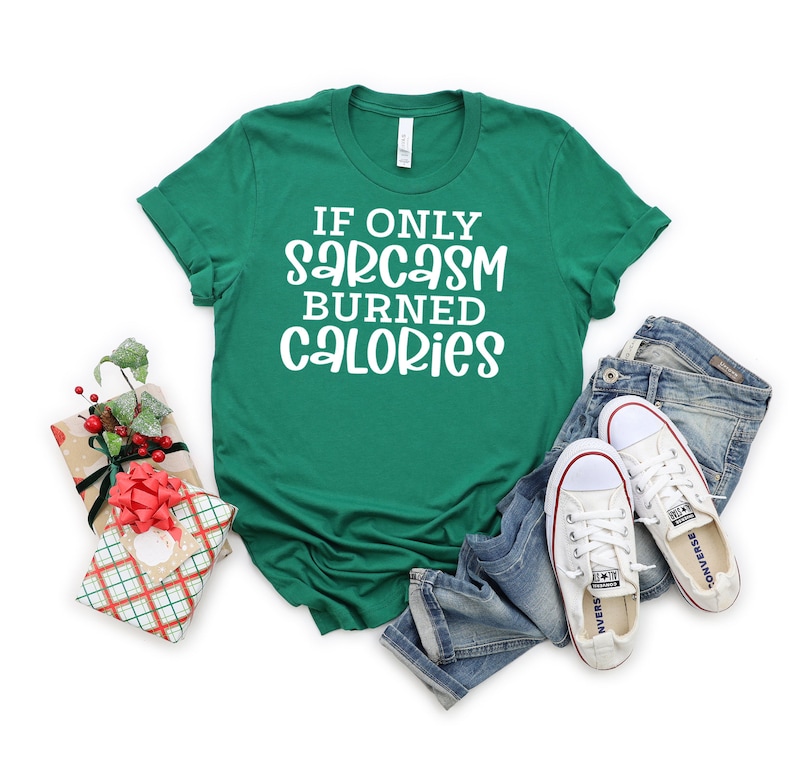 May include: A green t-shirt with white text that reads "If only sarcasm burned calories". The shirt is lying on a white background with a pair of blue jeans and white trainers.