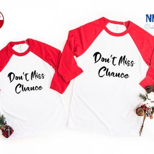 May include: Two white and red baseball shirts with the text "Don't Miss Chance" in black. The shirts are laid out on a white surface with a sprig of greenery and a red and white candy cane.
