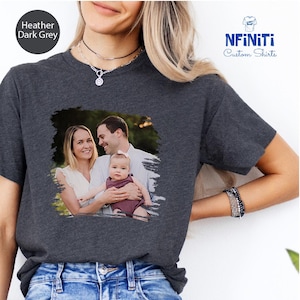 May include: A heather dark grey t-shirt with a photo of a family of three. The photo is printed on the shirt with a distressed, brushstroke effect. The family is standing in a grassy area with trees in the background. The text "NFINITI Custom Shirts" is printed in white on a black background in the upper right corner of the image.