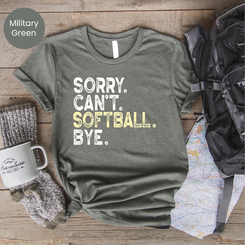 May include: Military green t-shirt with the text "SORRY. CAN'T. SOFTBALL. BYE." in white and yellow. A white mug with the text "Adventure Begins" and a black backpack are also visible.