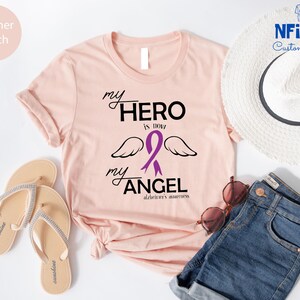 May include: Heather peach t-shirt with the text "my HERO is now my ANGEL" and a purple ribbon with wings, representing Alzheimer's awareness. The shirt is styled with a white hat, sunglasses, and denim shorts.