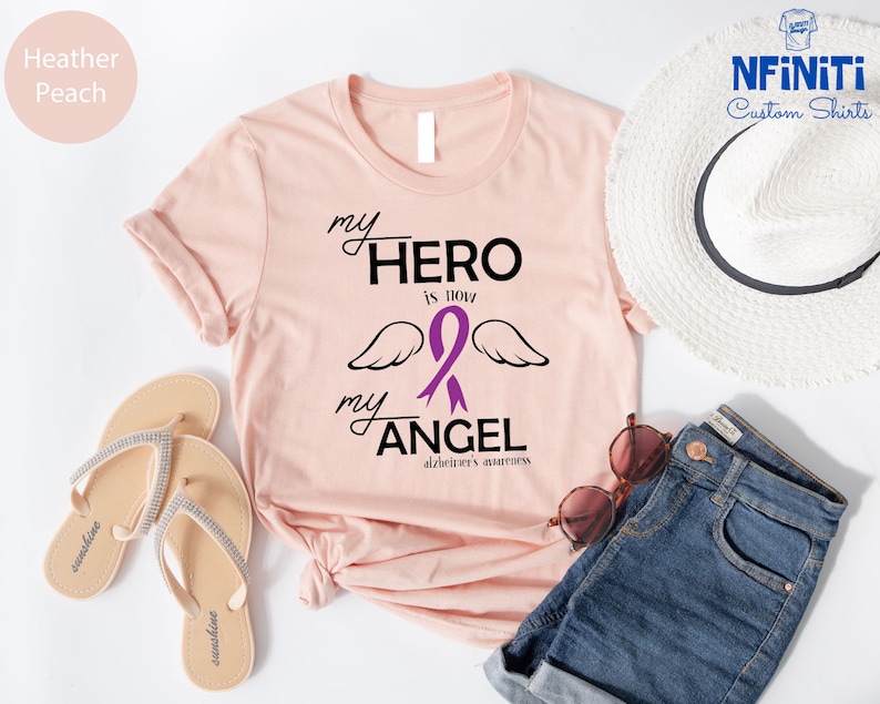 May include: Heather peach t-shirt with the text "my HERO is now my ANGEL" and a purple ribbon with wings, representing Alzheimer's awareness. The shirt is styled with a white hat, sunglasses, and denim shorts.