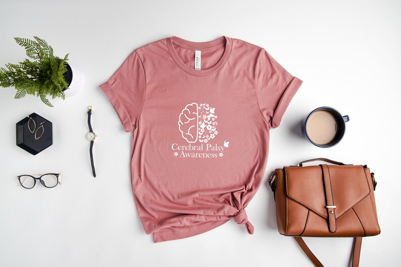 Może przedstawiać: R&oacute;żowy t-shirt z białym nadrukiem przedstawiającym m&oacute;zg z kwiatami i motylami. Pod nadrukiem znajduje się tekst "Cerebral Palsy Awareness".