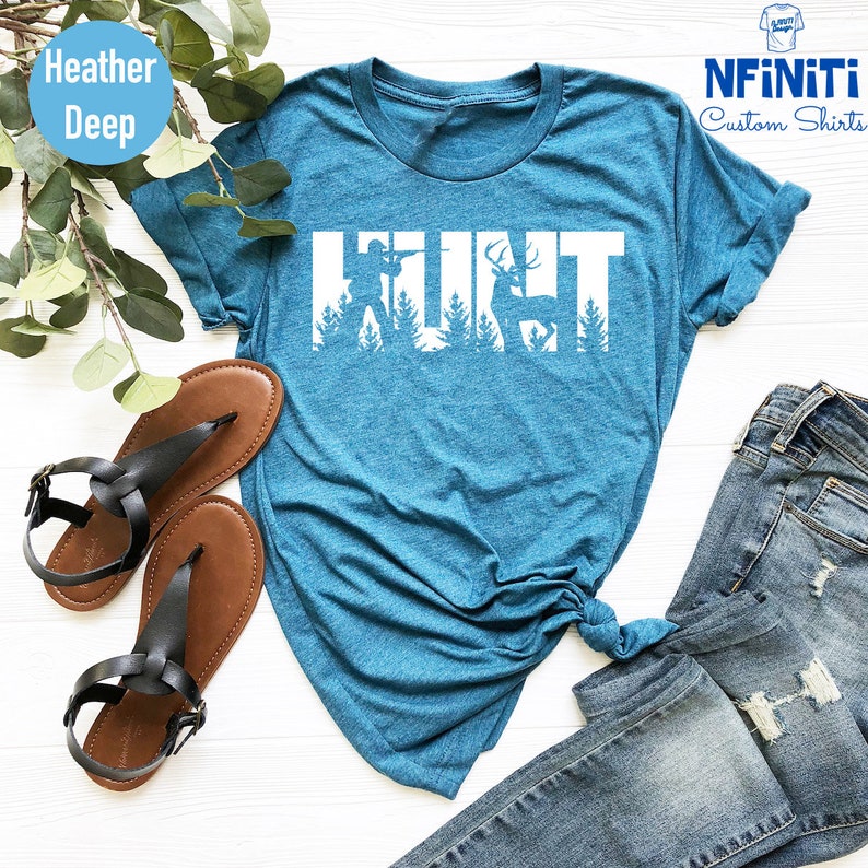 May include: A teal blue t-shirt with the word "HUNT" in white text. The letters are made up of silhouettes of a hunter, a deer, and trees. The shirt is laid out on a white surface with a pair of brown sandals and blue jeans.