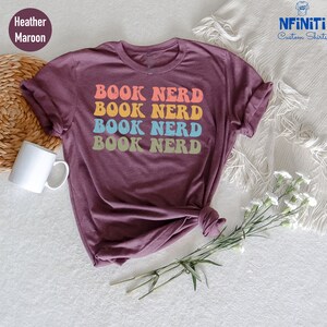 May include: A heather maroon t-shirt with the text "BOOK NERD" repeated four times in a retro rainbow font.