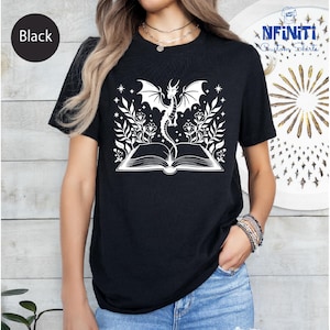 Op de afbeelding: Zwart T-shirt met een witte afbeelding van een draak die oprijst uit een open boek, omringd door bloemen en sterren. De tekst "NFINITI Custom Shirts" is zichtbaar. Het shirt heeft een ronde hals.