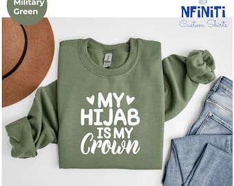 May include: A green sweatshirt with the text "My Hijab is my Crown" in white lettering. The sweatshirt is folded and laid out on a white surface with a brown hat and blue jeans in the background.