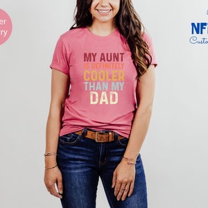 May include: A pink t-shirt with the text "My Aunt is Definitely Cooler Than My Dad" in a retro font with colorful letters.