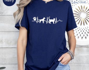 May include: Navy blue t-shirt with a white graphic of a dog paw, dog, and cat in a heartbeat line. The shirt has short sleeves and a crew neck. The word "Navy" is in a navy blue circle in the upper left corner.