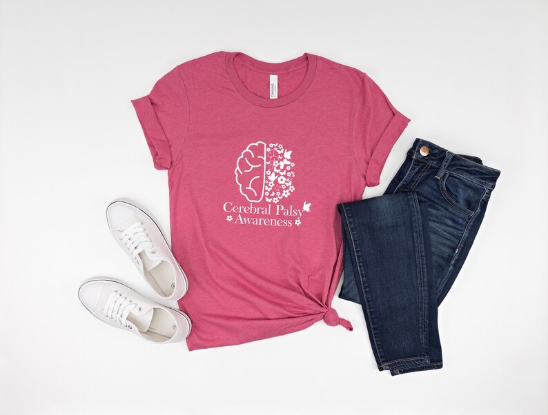 Może przedstawiać: R&oacute;żowy t-shirt z białym nadrukiem przedstawiającym m&oacute;zg z kwiatami i motylami. Pod nadrukiem znajduje się tekst "Cerebral Palsy Awareness". T-shirt jest zestawiony z niebieskimi dżinsami i białymi butami sportowymi.