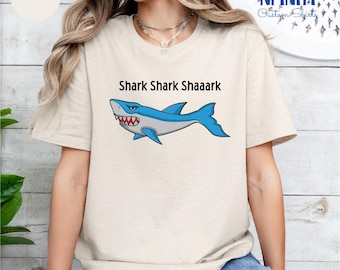 May include: A sand-colored t-shirt with a cartoon shark graphic and the text "Shark Shark Shaaark" printed on the front.