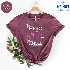 May include: Heather maroon t-shirt with the text "my HERO is now my ANGEL" in white script. A purple awareness ribbon and white wings are featured in the design. The phrase "alzheimer's awareness" is printed below the angel text.