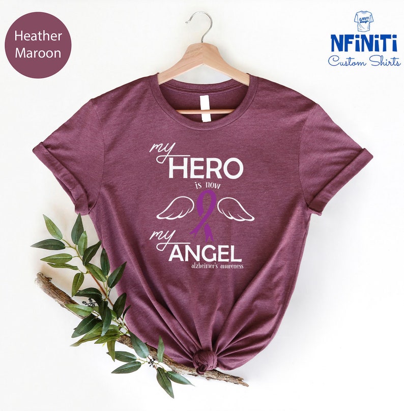 May include: Heather maroon t-shirt with the text "my HERO is now my ANGEL" in white script. A purple awareness ribbon and white wings are featured in the design. The phrase "alzheimer's awareness" is printed below the angel text.