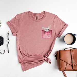 May include: Mauve t-shirt with a pocket design showing a watercolour illustration of a small, light-coloured animal. The shirt has rolled-up sleeves and a knotted hem. Accessories include a watch, glasses, and a brown handbag.