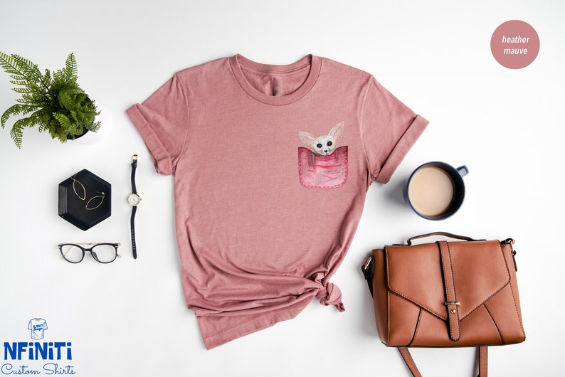 May include: Mauve t-shirt with a pocket design showing a watercolour illustration of a small, light-coloured animal. The shirt has rolled-up sleeves and a knotted hem. Accessories include a watch, glasses, and a brown handbag.