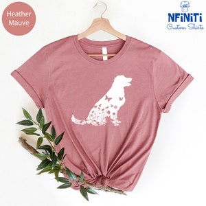 May include: Heather mauve t-shirt with a white silhouette of a dog with flowers and butterflies inside the silhouette. The shirt is on a hanger and has a white background.