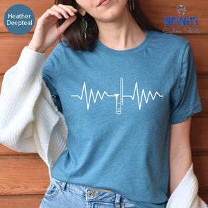 May include: A teal t-shirt with a white graphic of a trombone integrated into a heartbeat line. The shirt has short sleeves and is made of a soft material. The text "Heather Deepteal" is in the upper left corner.