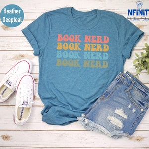 May include: A teal blue t-shirt with the text "BOOK NERD" repeated four times in a retro font. The text is in shades of orange, pink, yellow, and blue. The shirt is laid out on a wooden surface with a pair of white sneakers and denim shorts.