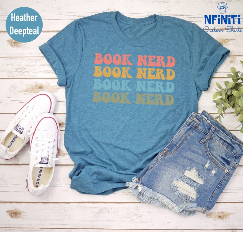 May include: A teal blue t-shirt with the text "BOOK NERD" repeated four times in a retro font. The text is in shades of orange, pink, yellow, and blue. The shirt is laid out on a wooden surface with a pair of white sneakers and denim shorts.