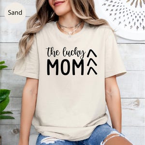 May include: Sand-colored t-shirt with the text "the lucky MOM" in black. The text is in a handwritten style font. The shirt has short sleeves and a crew neck. The model is wearing a necklace and blue jeans.