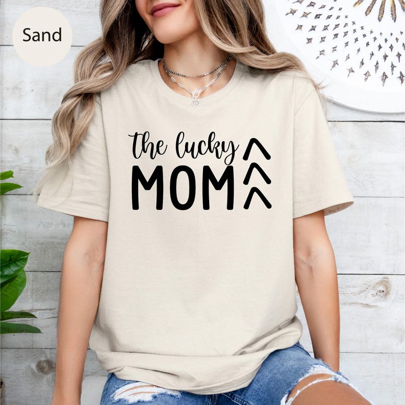May include: Sand-colored t-shirt with the text "the lucky MOM" in black. The text is in a handwritten style font. The shirt has short sleeves and a crew neck. The model is wearing a necklace and blue jeans.