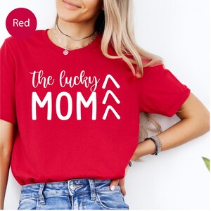 May include: A red t-shirt with the text "the lucky MOM" in white script and block letters. Above the word "MOM" are two upward-pointing arrows. The shirt is short-sleeved and made of a soft material.