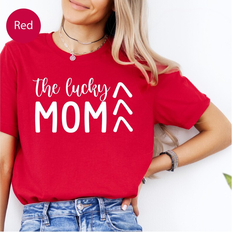 May include: A red t-shirt with the text "the lucky MOM" in white script and block letters. Above the word "MOM" are two upward-pointing arrows. The shirt is short-sleeved and made of a soft material.