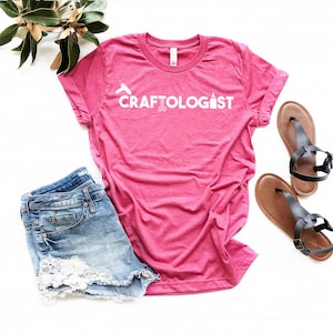 May include: A pink t-shirt with the text "CRAFTOLOGIST" in white. The text is accompanied by a graphic of a glue bottle, scissors, and a paintbrush. The shirt is laying on a white surface with a pair of brown sandals and denim shorts with lace trim.