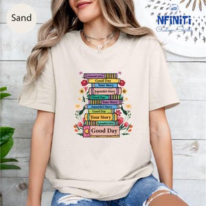 May include: A white t-shirt with a stack of colorful books with the text "Amanda's Story", "Good Day", and "Your Story" repeated on each book. The books are surrounded by floral illustrations.