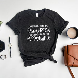 May include: A dark gray t-shirt with the text "You people must be exhausted from watching me do everything" in white lettering.