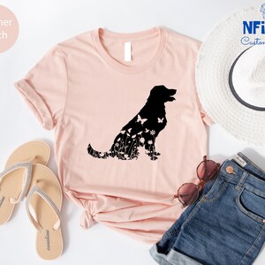 May include: A heather peach t-shirt with a black silhouette of a dog made of flowers and butterflies. The shirt is laid out on a white surface with a pair of sandals, a straw hat, and denim shorts.