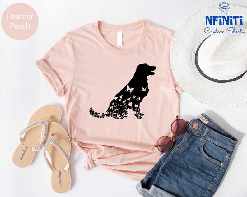 May include: A heather peach t-shirt with a black silhouette of a dog made of flowers and butterflies. The shirt is laid out on a white surface with a pair of sandals, a straw hat, and denim shorts.