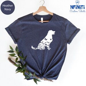 May include: A heather navy blue t-shirt with a white silhouette of a dog with flowers and butterflies inside the outline. The shirt is on a hanger.