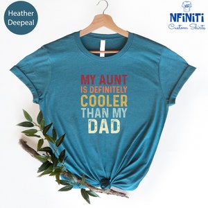 May include: Teal blue t-shirt with the text "My Aunt is Definitely Cooler Than My Dad" in a vintage style font with red, orange, and yellow colors.
