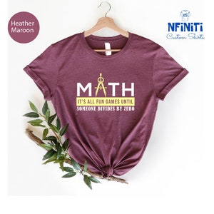 Op de afbeelding: Heather maroon t-shirt met een witte afbeelding met de tekst "MATH" met een passer en een gradenboog. De tekst eronder zegt "It's all fun games until someone divides by zero."