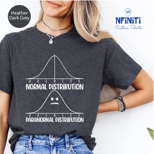 May include: A heather dark grey t-shirt with a white graphic of two bell curves. The top curve is labeled "Normal Distribution" and the bottom curve is labeled "Paranormal Distribution".
