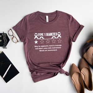 K&ouml;nnte beinhalten: Ein T-Shirt in Heidelbeer-Maroon mit einer wei&szlig;en Grafik eines Bandes und dem Text "TYPE 1 DIABETES" mit einer Ein-Sterne-Bewertung und dem Text "Way too expensive, hard to manage, And didn't come with instructions Would not recommend."