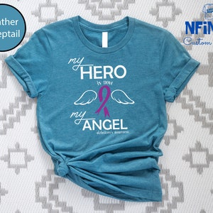May include: A teal t-shirt with the text "my HERO is now my ANGEL" in white, with a purple ribbon and angel wings. The words "Alzheimer's awareness" are below the text. The shirt is from NFiniTi Custom Shirts.