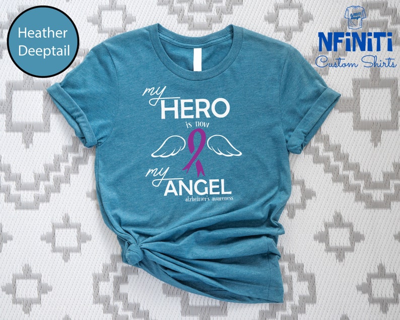 May include: A teal t-shirt with the text "my HERO is now my ANGEL" in white, with a purple ribbon and angel wings. The words "Alzheimer's awareness" are below the text. The shirt is from NFiniTi Custom Shirts.