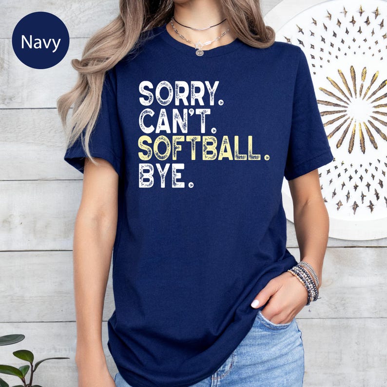 May include: Navy blue t-shirt with the text "SORRY. CAN'T. SOFTBALL. BYE." in white and yellow lettering. The shirt has short sleeves and a crew neck. The word "Navy" is in a navy blue circle in the upper left corner.