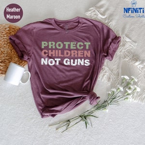 May include: A heather maroon t-shirt with the text "PROTECT CHILDREN NOT GUNS" in white letters with a green, pink, and orange distressed effect.