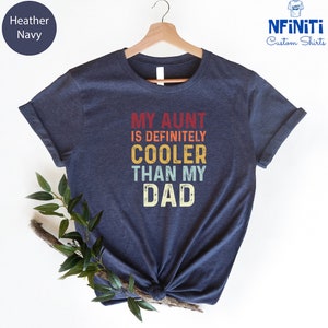 May include: A heather navy blue t-shirt with the text "My Aunt is Definitely Cooler Than My Dad" in a vintage style font with colorful accents.