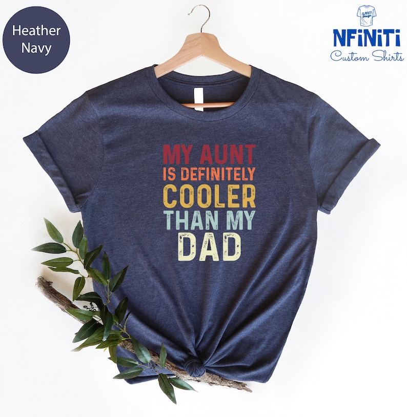 May include: A heather navy blue t-shirt with the text "My Aunt is Definitely Cooler Than My Dad" in a vintage style font with colorful accents.